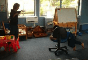 A child in a classroom with their body across an chair with wheels, pushing the chair with their legs and 'chasing' their teacher