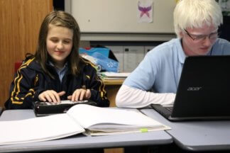 Brianna and Brayden sitting at desks with a laptop, Braille Sense U2 and hard copy braille