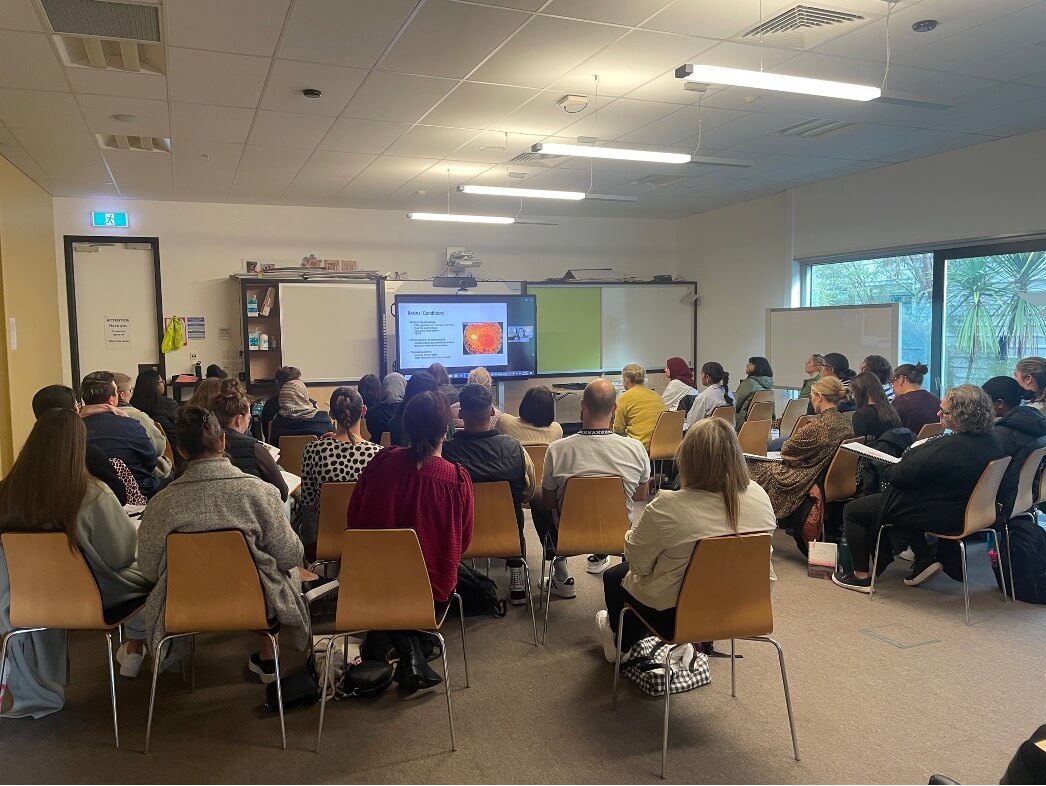 Figure 1 - A large group of teachers & teacher aides, seated in rows of chairs, watching a presentation on a large screen about various eye conditions