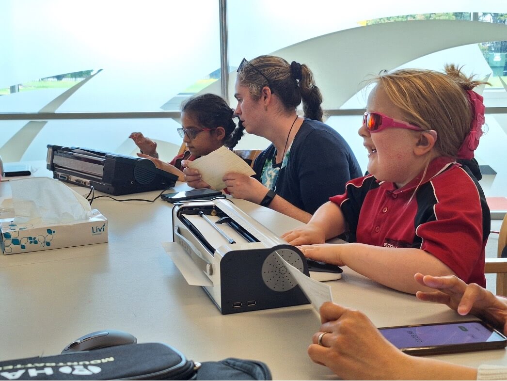 Figure 2 - One female Resource Teacher Vision sitting between two young girls who are typing on their braille device